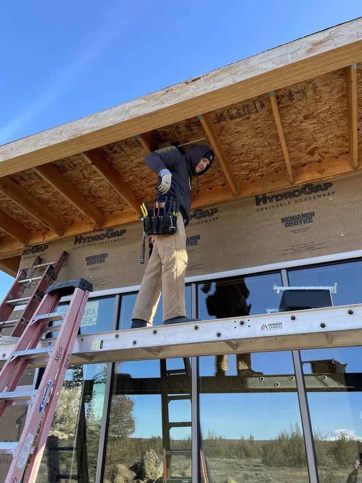 An electrician on a plank between two ladders, working at the eaves of a new home with tall windows