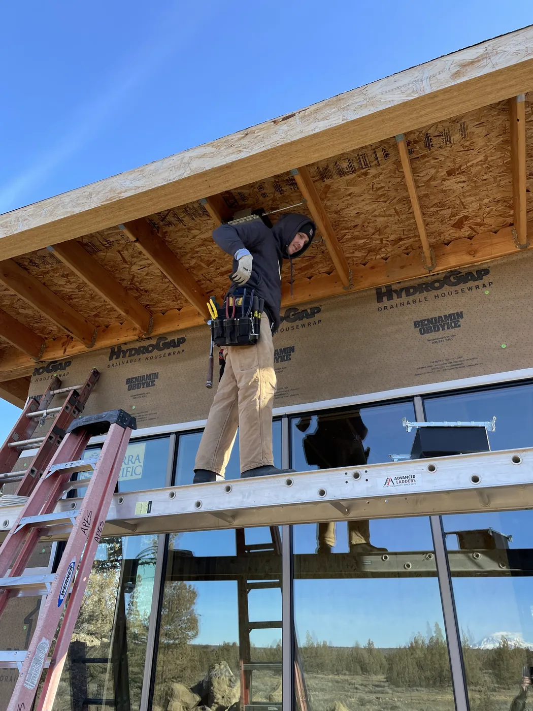 An electrician on a plank between two ladders, working at the eaves of a new home with tall windows